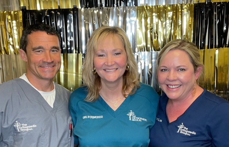 From left to right - Dr. Spain is pictured in gray scrubs with Robin, RT, in teal scrubs, and another employee of The Orthopaedic Group in navy blue scrubs. They are smiling and standing in front of a shiny black, gold, and silver backdrop.