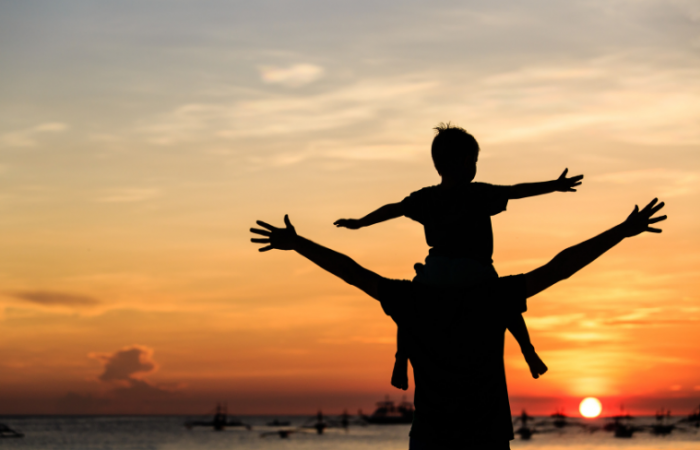 a man with a young boy on his shoulders with arms out at sunset